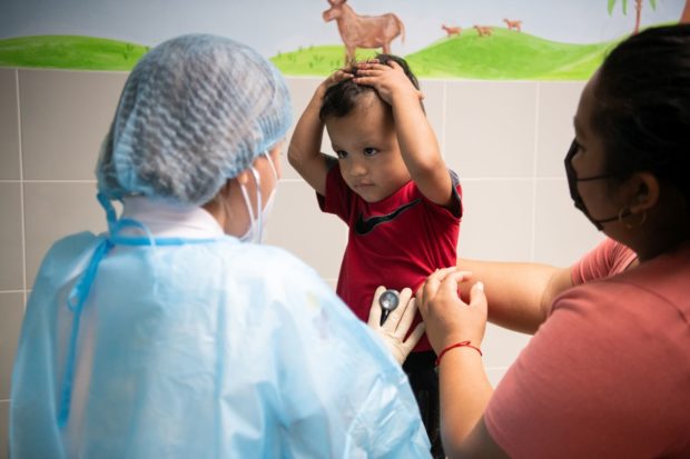 Doctor in blue medical gown and mask with back to us holds stethoscope to toddler in red who is standing with both hands on his head. His mother on the right side holds onto her son with her right arm