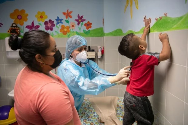 The doctor in blue medical garb with mask holds stethoscope to toddler's back. The boy in red points at a donkey in a mural on the wall. His mother in black mask and salmon top watches. them.