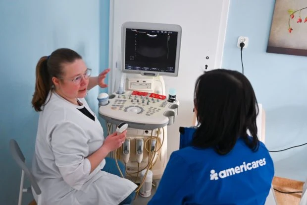 Health worker in white lab coat demonstrates an ultrasound machine showing monitor and controls to Americares staff member in blue t-shirt.