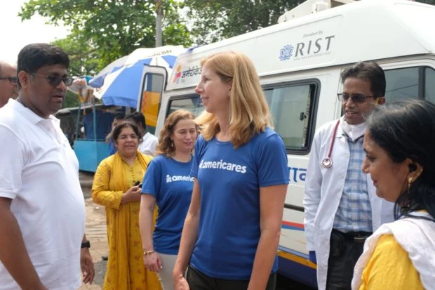 Americares CEO in blue americares shirt at the center of a group of staff members beside one of the Mobile Health Centers.