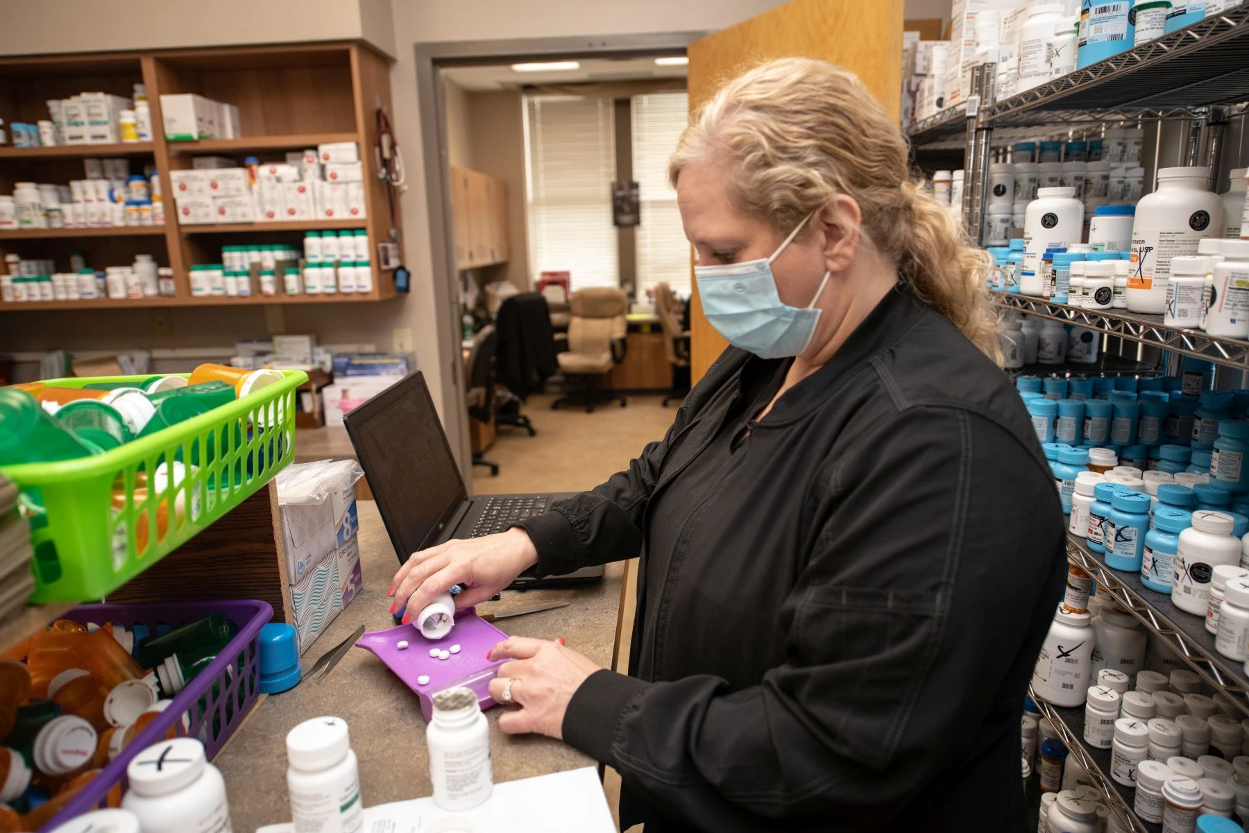 Pharmacist in black counting out pills next to a laptop computer with filled pharmacy shelves in the background