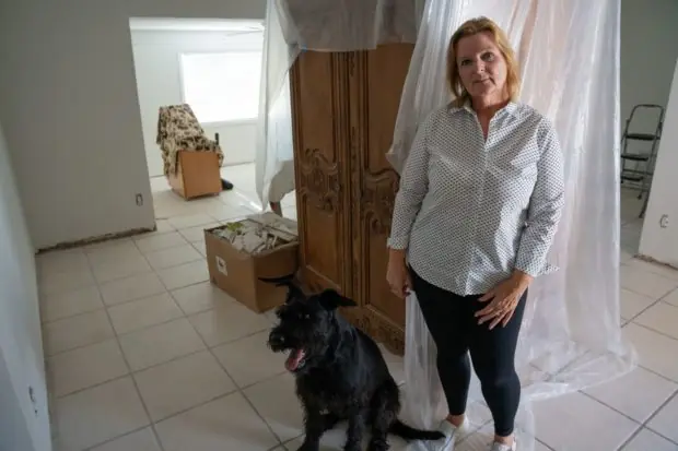 Woman with her black dog standing in front of a partially covered wooden wardrobe with two boxes of debris in background and evidence of damage along the baseboards.