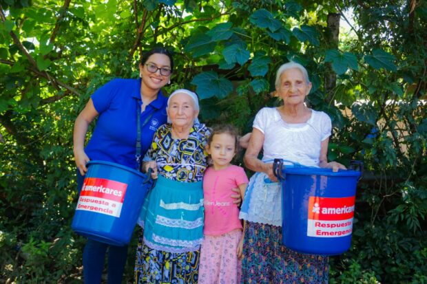 Americares team member in blue shirt distributing bucket hygiene kits to three family members - two elderly and one child.
