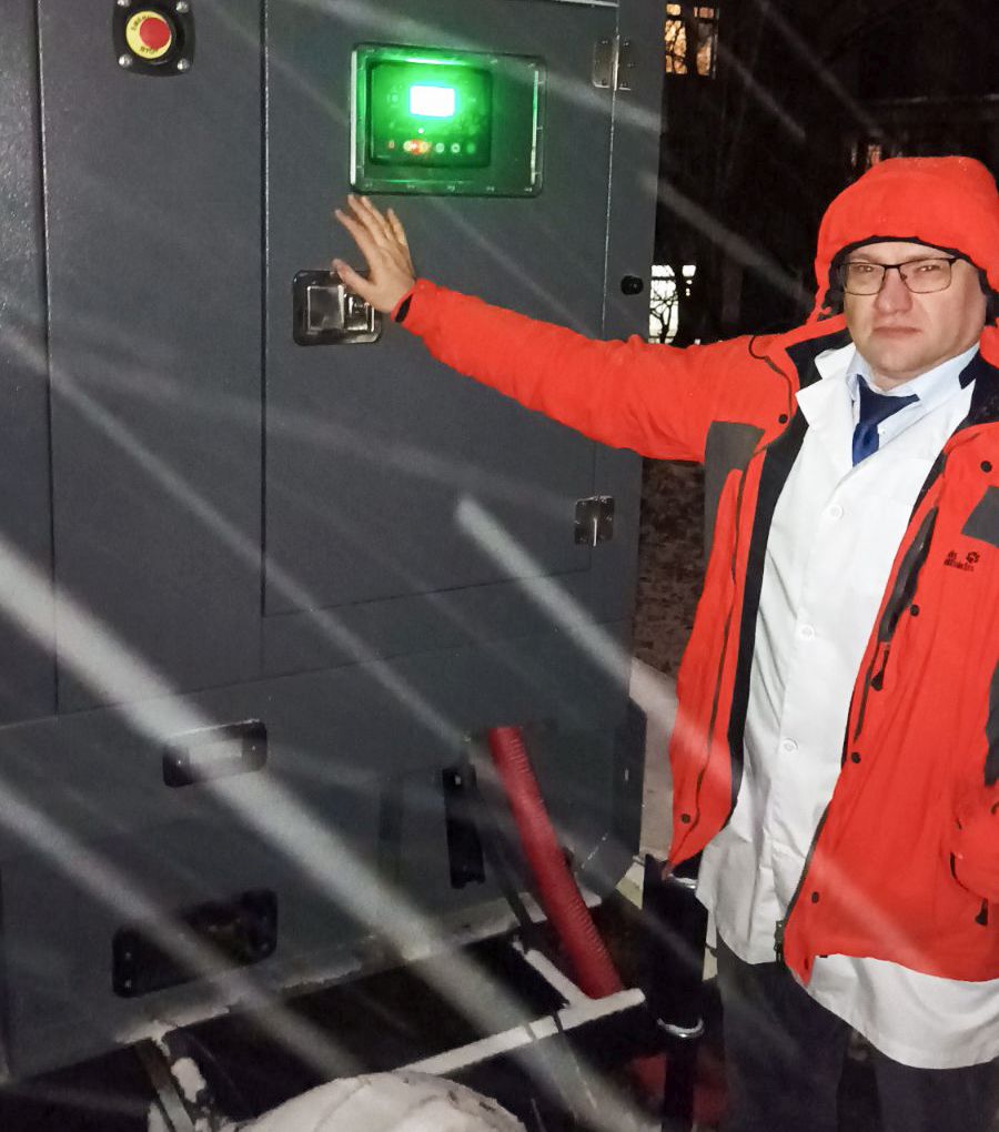 man in white lab coat under an orange jacket with hand on new installed generator with green light on in panel.