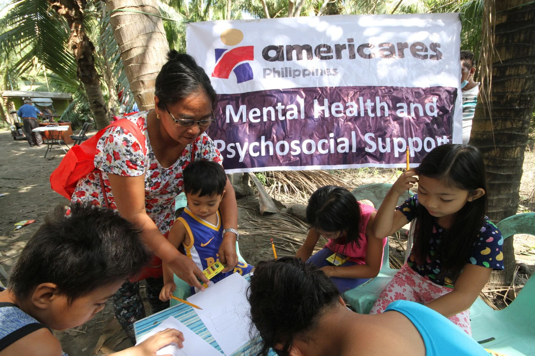 Health worker behind child in a circle of 5 children working with paper and pencils, drawing pictures in front of a sign that reads, "Americares Philippines, Mental Health and Psychosocial Support.
