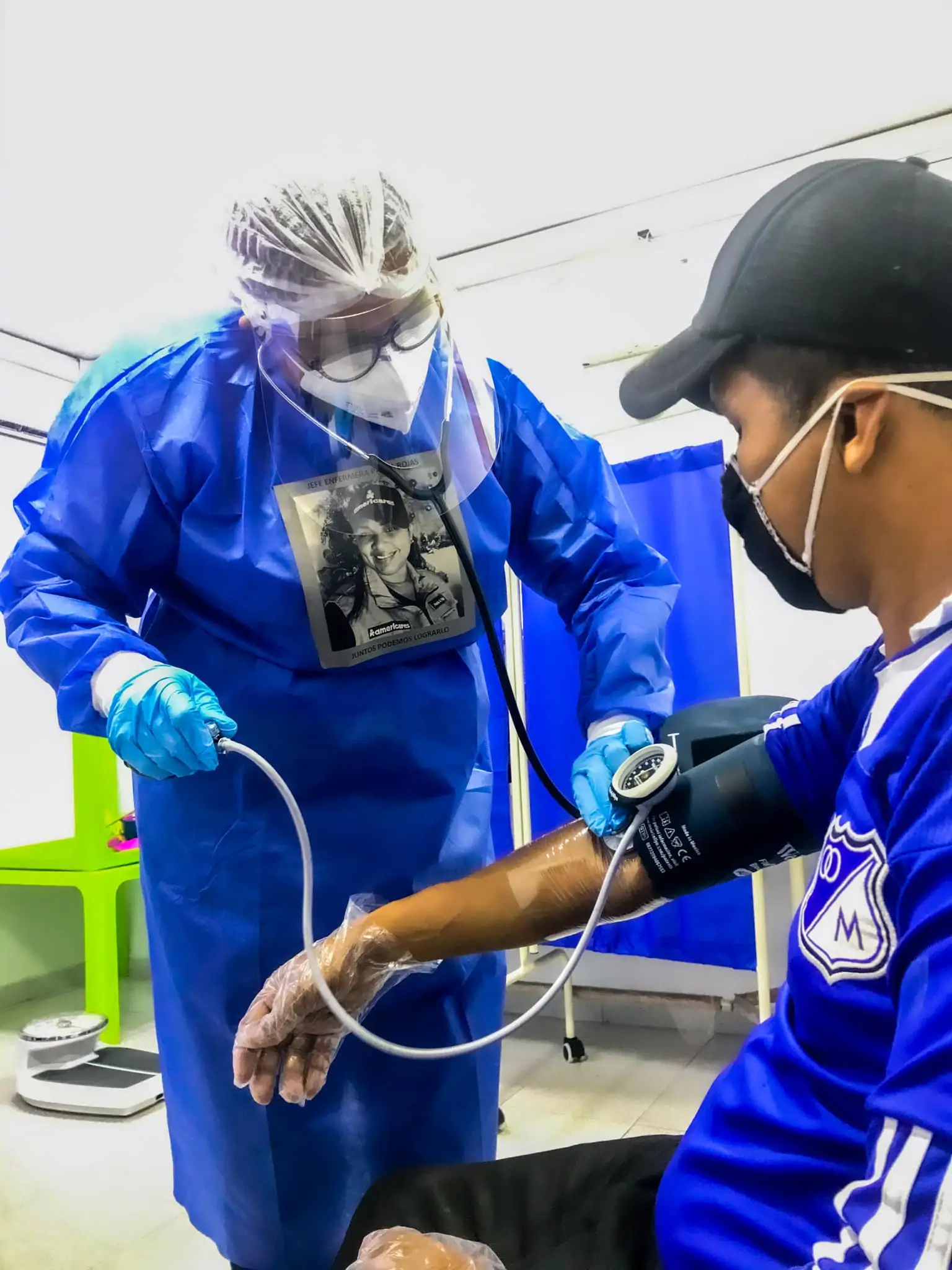 Clinic health worker in full PPE gear and nameplate checks the blood pressure of a patient wearing a mask and baseball cap.