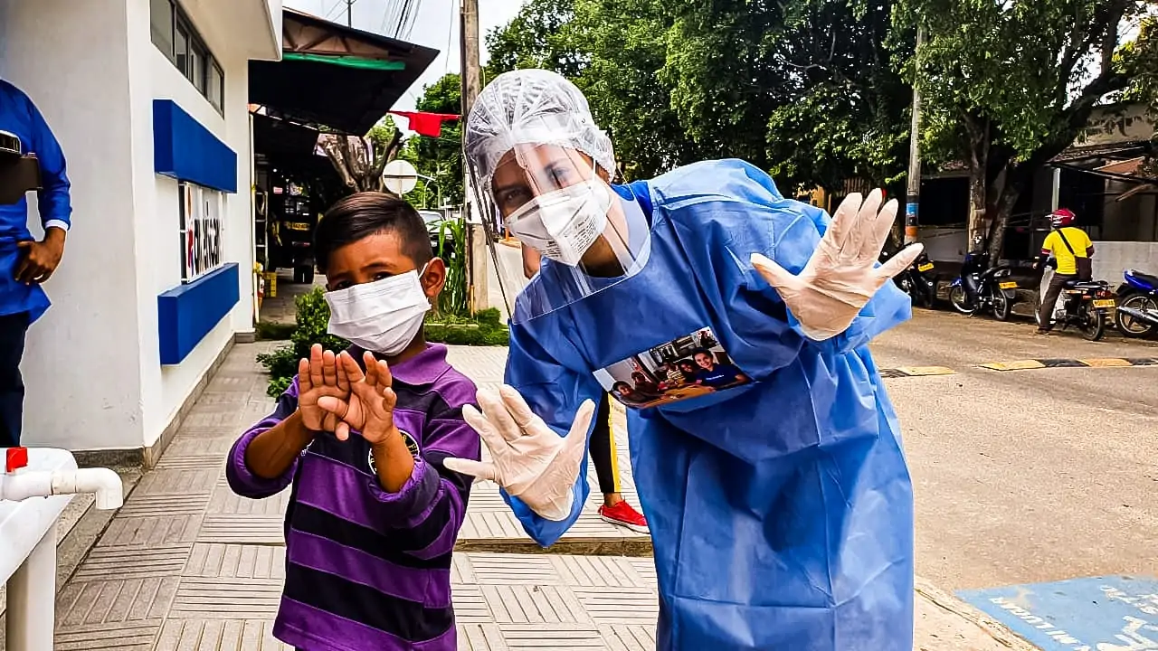 Healthcare worker in surgical mask and gown holds up her gloved hands with a child wearing a mask showing they washed their hands.