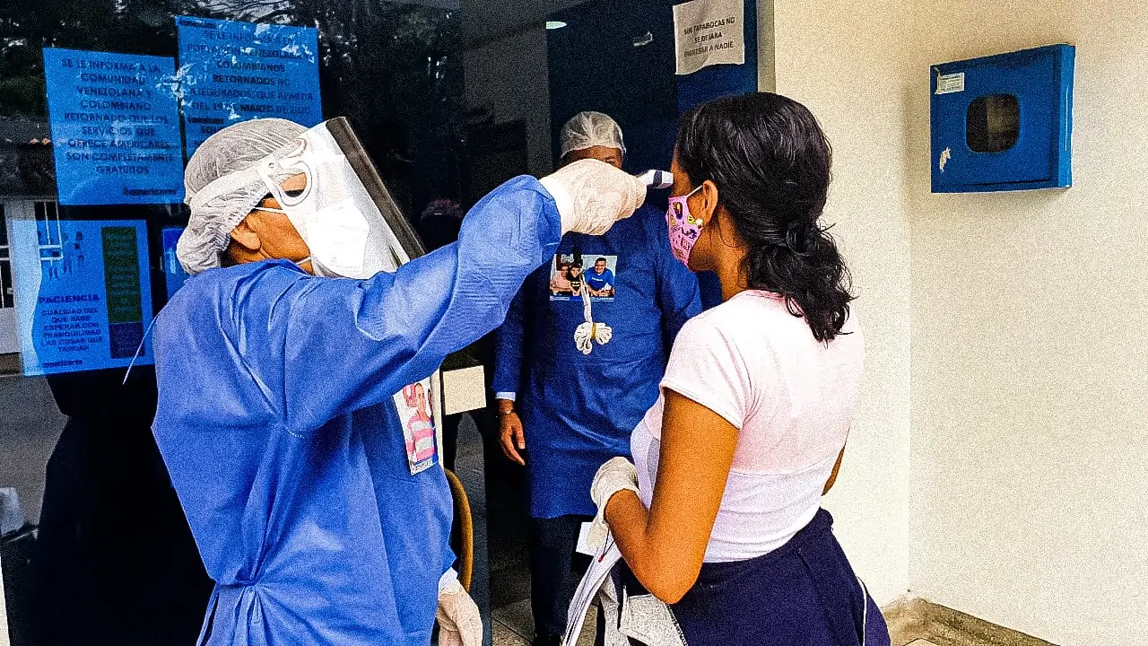 A woman in a face masks gets her temperature taken by two medical workers in full PPE gear.