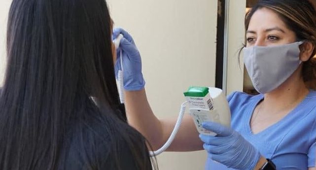 Image of clinic staff member wearing a mask and taking a young woman's temperature.