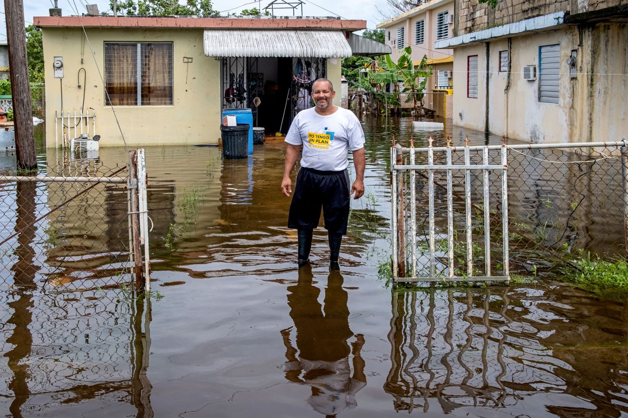 Man in white t-shirt and black shorts stands knee deep in floodwaters in front of his home in Puerto Rico