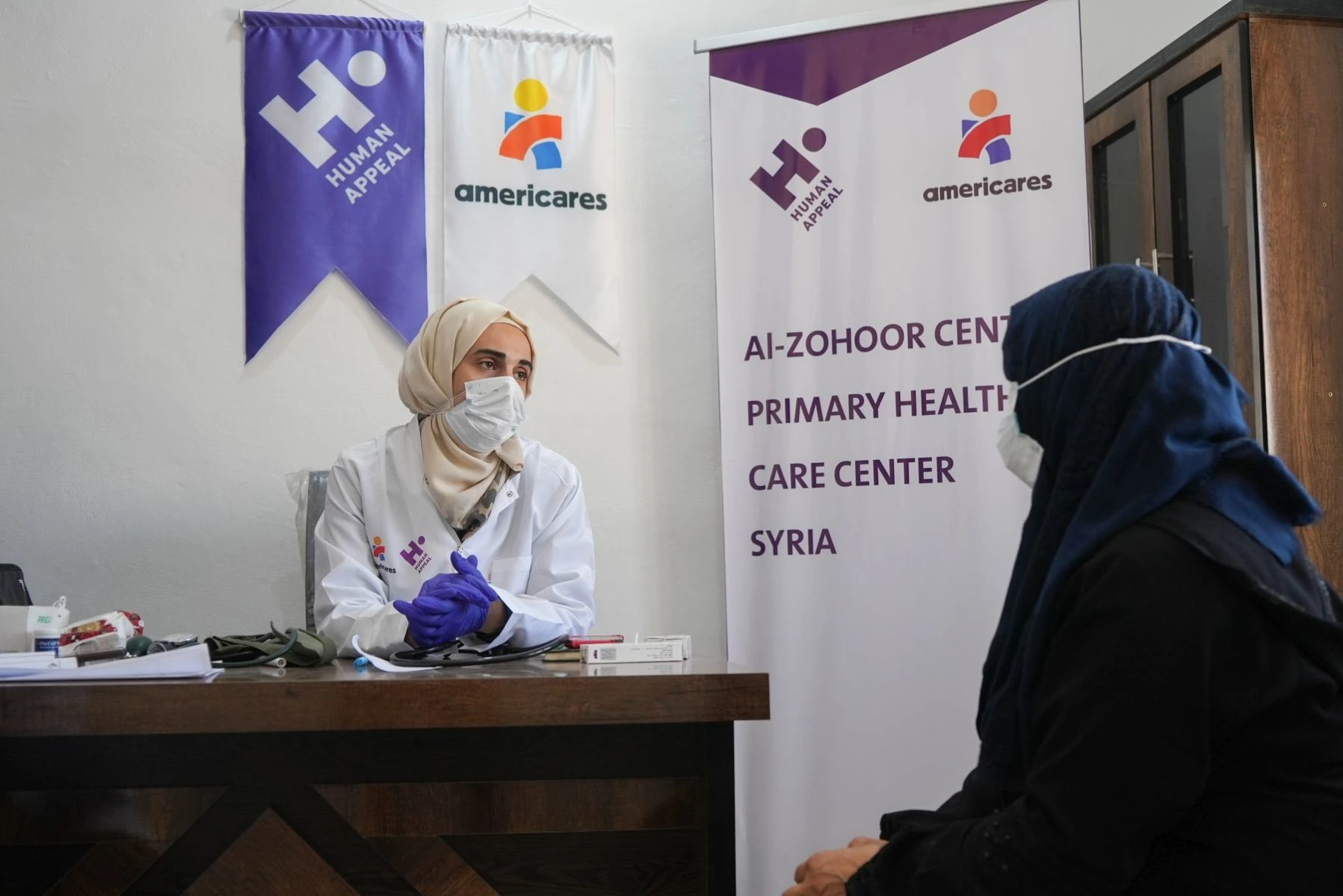 Health worker in mask, lab coat and hijab with gloves speaks with patient seated in mask and hijab. Human Appeal and Americares signage behind health worker