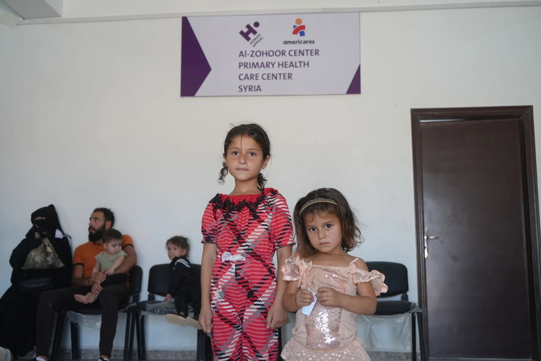 Two young girls, one on left taller in bright pink outfit, younger one on right in light pink. Family with children seated in the background. Sign with Human Appeal, Americares and health center name on wall.