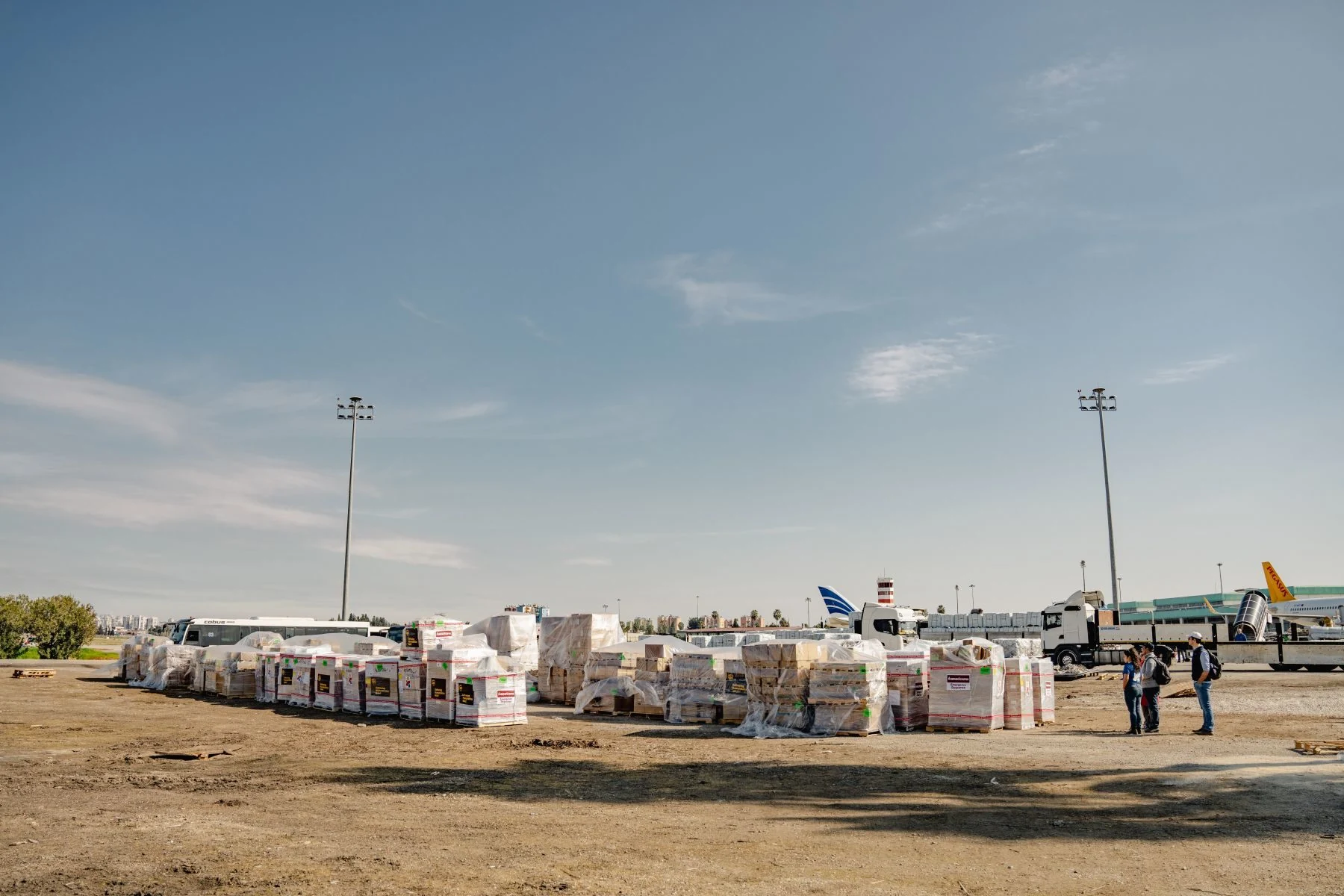 A long camera shot of a large shipment of relief supplies that sits at the airport after being unloaded with three team members checking the shipment at the right. Planes visible in the background.