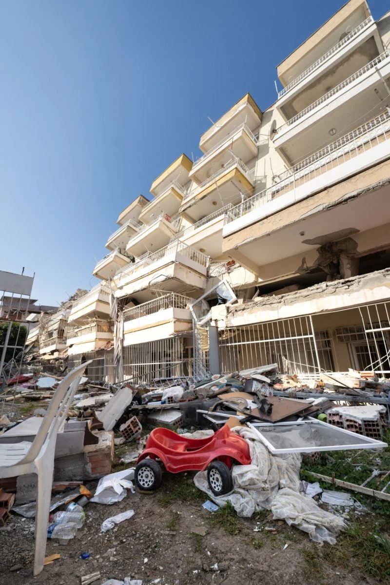 a child's red toy car sits in foreground in sea of contents spread on ground from the ruined apartment buildings behind.