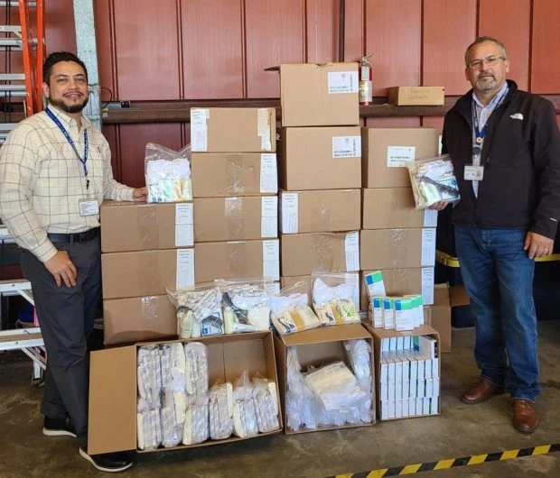 One man stands to left of stacked open and unopened boxes of medical supplies. A second man in dark jacket stands to the right side holding a clear package of supplies.