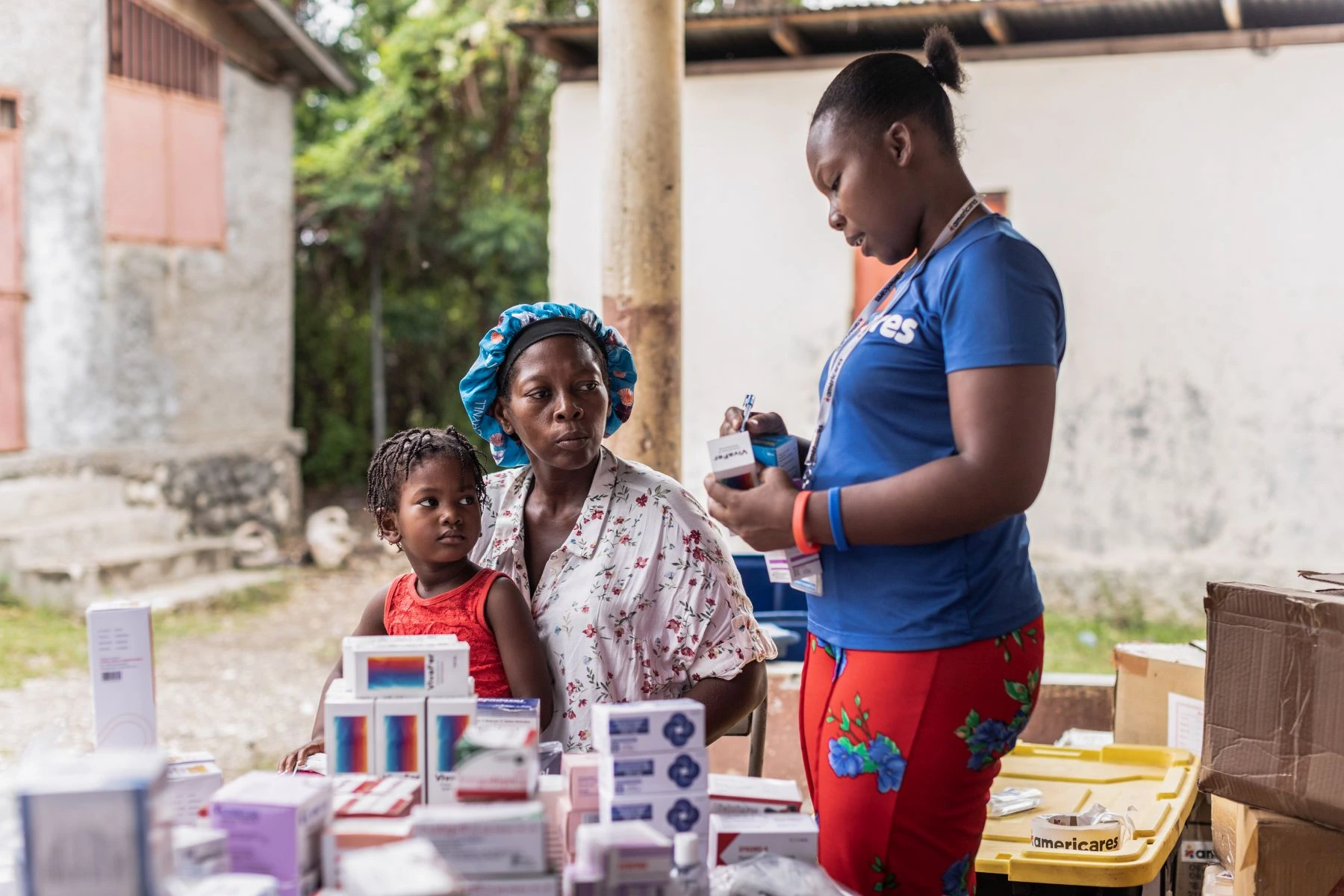 Standing medical team member in blue shirt prepares medicine for mother and daughter seated at table with supplies.