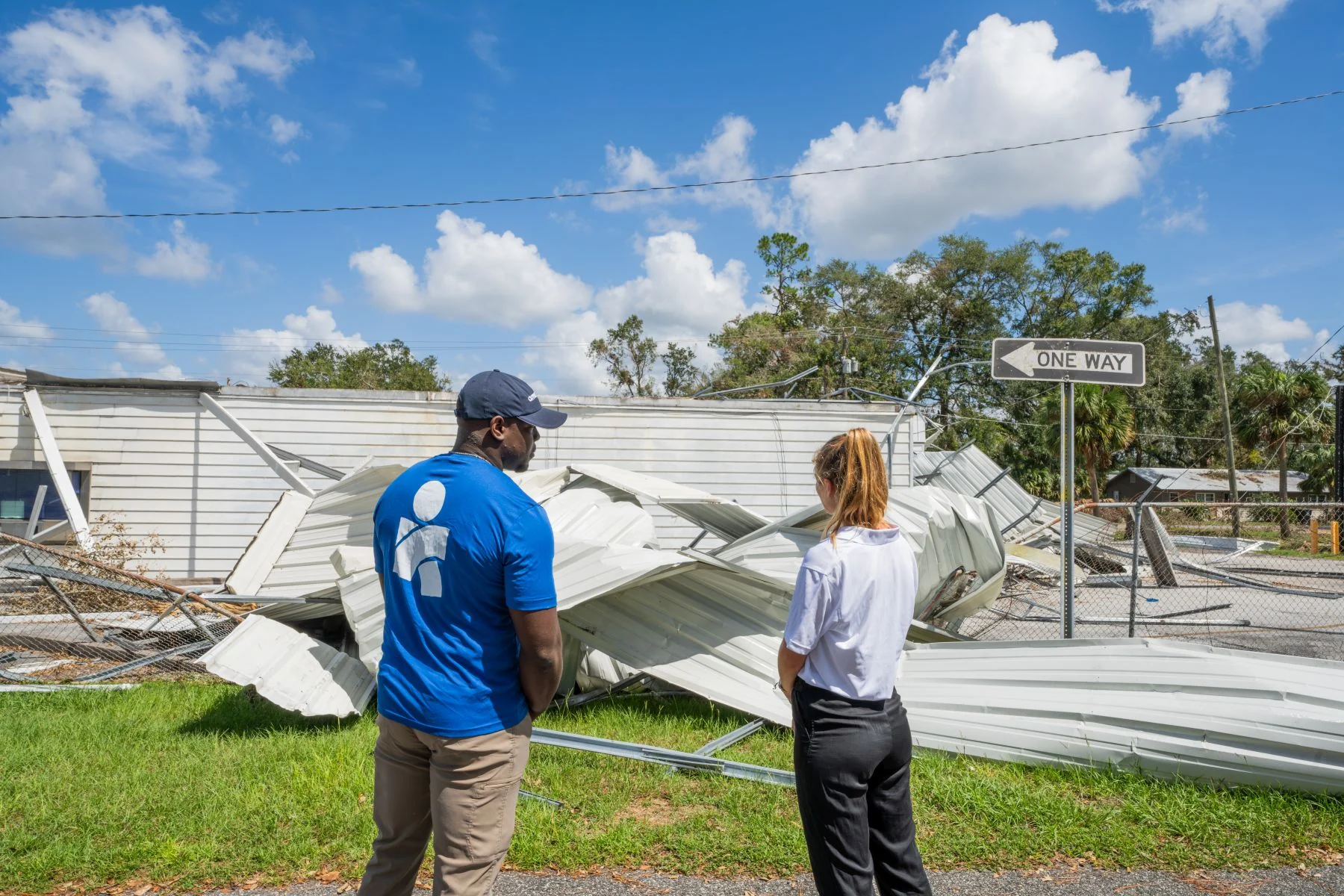 Americares team member in blue shirt stands beside person in white shirt surveying a destroyed building with one way sign still standing in wreckage.
