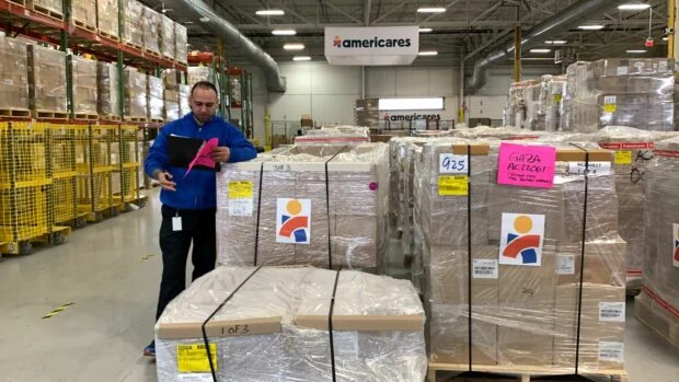 Prepared pallets of relief supplies with Americares logo and Gaza sign are being checked by staff member in blue jersey.