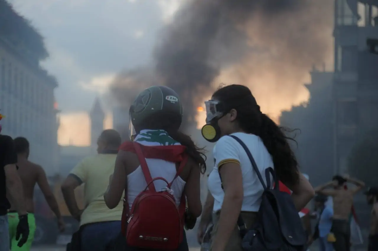 Two women in a small crowd protect themselves from smoke with a dust respirator and googles with a city building burning in the background.