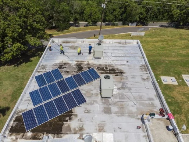 Solar panels being installed on roof of clinic with two installers in yellow vests and one americares staff in blue shirt visible in drone view