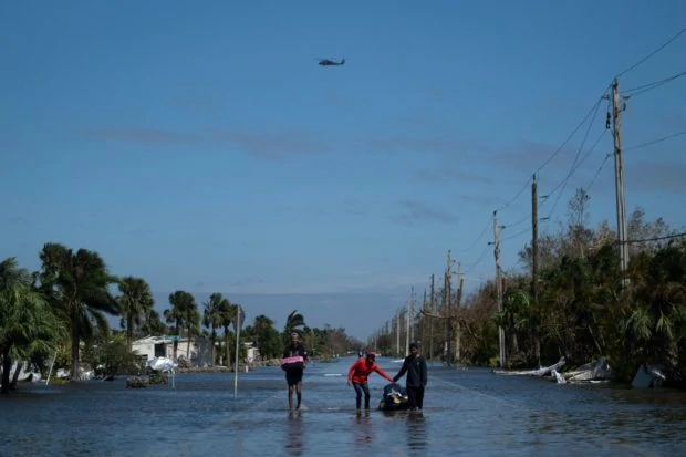 Three people carrying belongings in flooded street with utility poles on right side and flooded homes and palm trees on the left side.