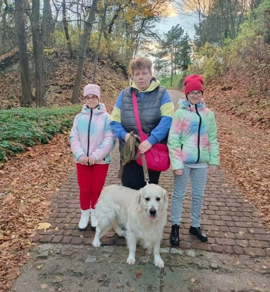 Oksana stands with her two daughters on either side and their white dog in front on a leash. They stand on a cobblestone path in a wooded area in winter. Ground is covered in leaves.