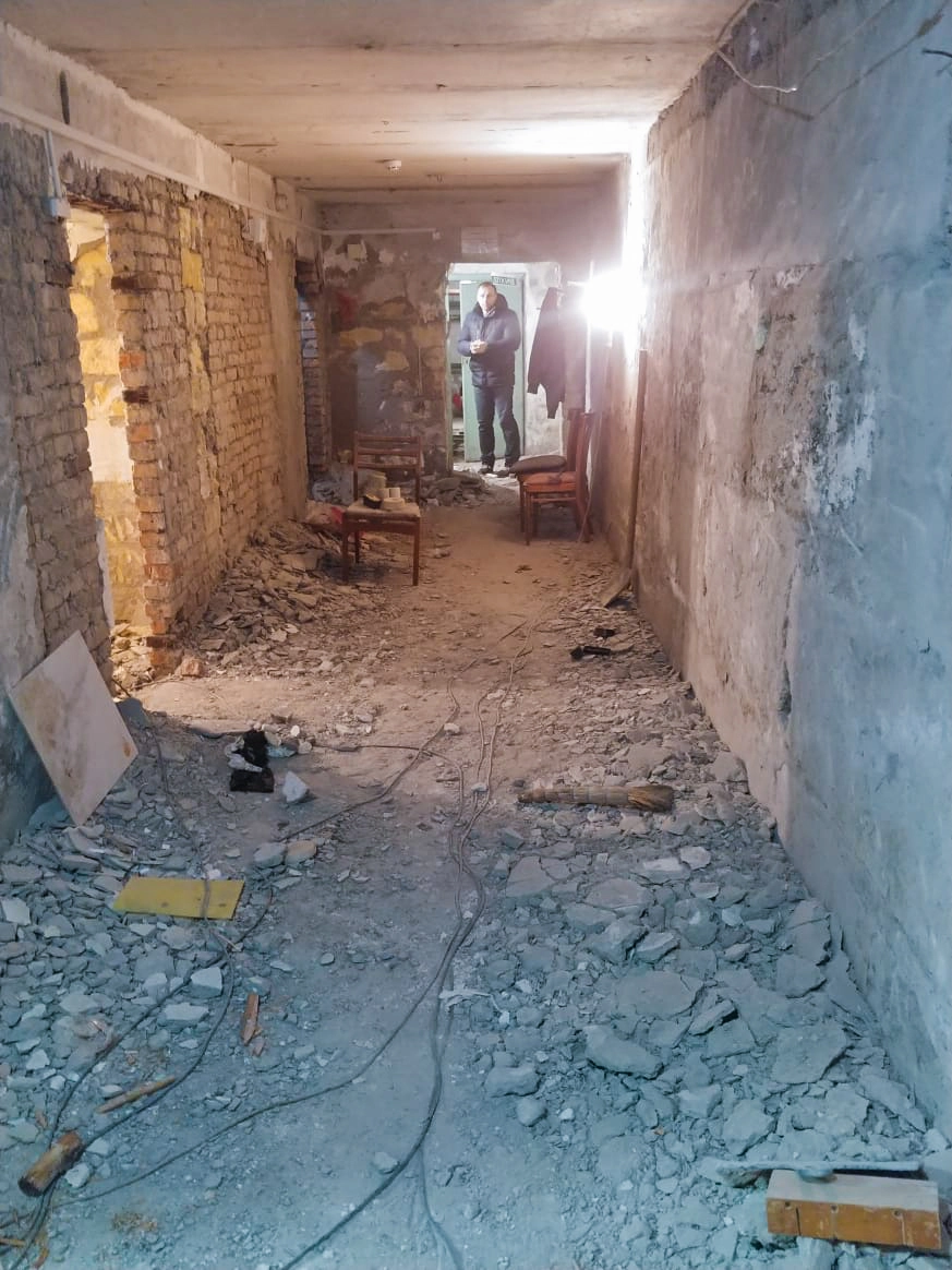 A long narrow basement room with exposed brick and concrete debris and one door on left and one at the far end with man standing in the door opening