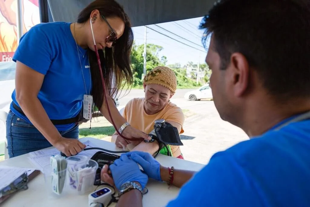 Two Americares medical staff take the blood pressure and heart rate of an older woman.