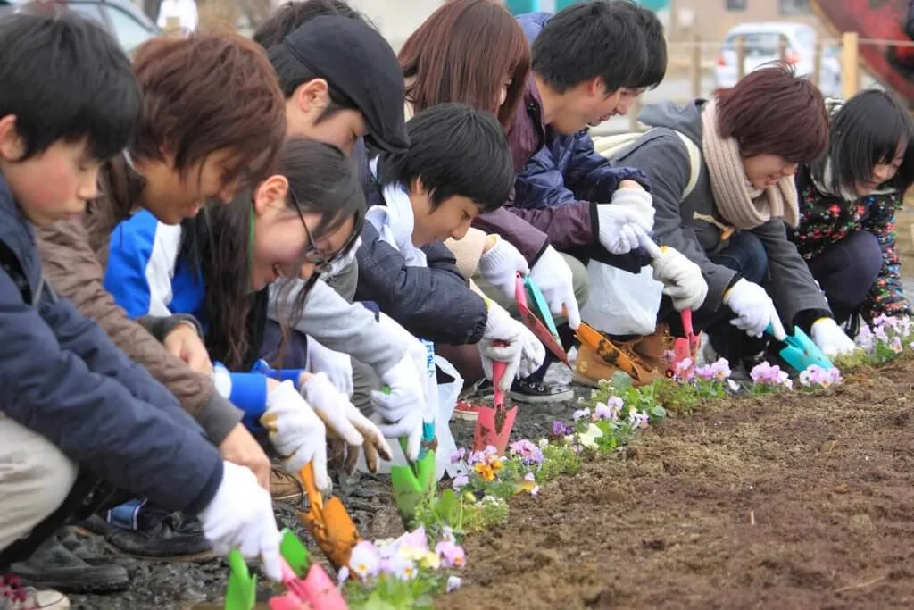 A line of young children plant flowers with plastic shovels and smiles.