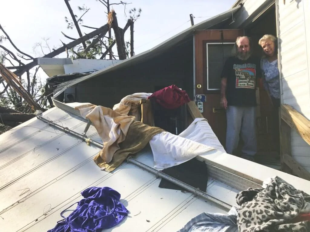 A husband and wife standing in their doorway of their damaged home with clothing and towels draping their staircase and fallen roof.
